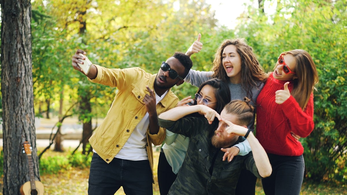 Playful youth men and women are taking selfie in park using smartphone, making funny faces and wearing sunglasses standing outdoors and laughing. Fun and autumn nature concept.